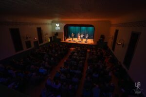 Five-Piece Band Performance Indoor theater performance with a five-member band on stage, audience seated, room full.