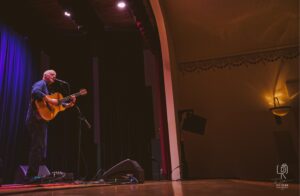 Guitarist on Dimly Lit Stage Musician performing on stage with guitar and microphone, purple curtain backdrop, dim lighting.