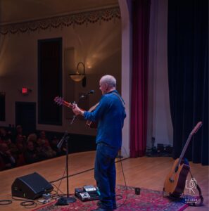 Acoustic Performance on Stage Musician on stage with guitar, microphone, wooden floor, red curtains, audience, audio equipment.