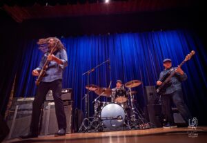 Energetic Trio Live Performance Musical trio on stage with guitarist, drummer, and bassist in front of blue curtain backdrop.