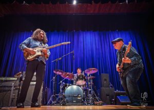 Stage Trio Performance Three-member band performing with guitar, bass, and drums on stage with blue curtains.
