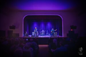 Purple-Lit Band Performance Band performing on stage with purple lighting, audience seated, dark curtains backdrop.