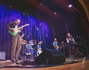 Indoor Stage Band Performance Musical performance on indoor stage with guitarist, keyboardist, saxophonist, drummer, and lights.