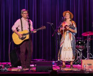 Stage Duo Performing Live man and woman performing on stage with instruments against purple curtain backdrop
