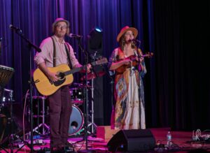 Acoustic Duo on Stage Musicians perform on stage with instruments, purple curtain backdrop, and stage lighting.