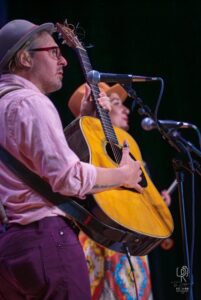 Acoustic Guitar Performance on Stage Musician in a hat and glasses singing with a guitar on stage; another person is partially visible.