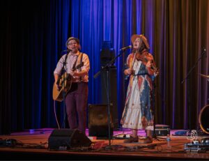 Stage Duo with Guitar and Violin Man and woman perform on stage with guitar and violin, colorful lighting, cables visible.