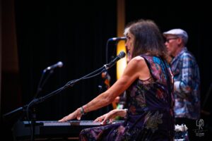 Keyboard Performance in Floral Dress Woman in floral dress playing keyboard on stage, near microphone, with person in background.