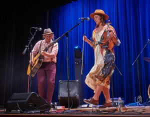 Acoustic Duo Performs Live on Stage Musicians playing guitar and violin on a stage with blue lighting and audio equipment nearby.