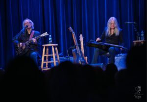 Stage Performance with Guitars Musicians seated on stage with guitars, blue curtains, and sound equipment in the background.