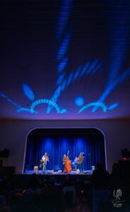 Jazz Ensemble in Vibrant Lighting Band performing on stage with blue and purple lighting, audience seated in the foreground.