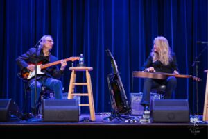 Guitar Duo on Stage Musicians on stage with blue curtains, playing electric and lap steel guitars; stool with water.