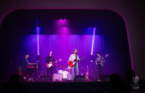 Stage Performance with Purple and Blue Lighting Five musicians perform on stage with purple lighting and various instruments visible.