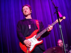 Electric Guitar Performance Under Purple Lights Musician playing red electric guitar on stage with purple lighting, drummer in background.