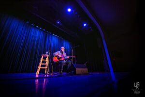 Acoustic Stage Performance Musician performing with guitar on blue-lit stage, mic and stool with water bottle nearby.