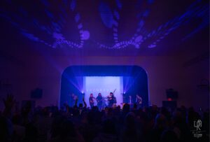 Live Concert in Vibrant Lights Live music show with purple-blue lights, indoor venue, musicians on stage, large audience present.