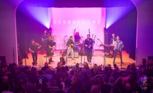 Skerryvore Live Concert Performance Band performs on stage with instruments and pink-purple lights; audience present.