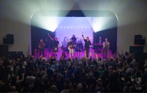 Energetic Live Concert Performance Band performing on stage with guitar, drums, violin, accordion; lively crowd engaging enthusiastically.