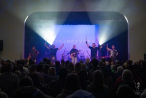 Skerryvore Band Live Performance Band performing with guitar, accordion, violin; "SKERRYVORE" on backdrop, engaged audience.