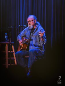 Acoustic Performance Under Warm Lights Man with beard and glasses plays acoustic guitar on stage, wearing denim, with microphone nearby.