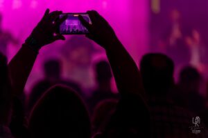 Concert Capture in Purple Hues Audience in purple light, person filming stage performance with phone showing performers.
