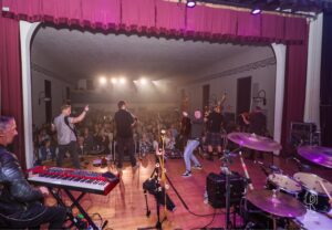 Energetic Band Performance in Theatrical Venue Band performs on stage in an indoor venue with engaged audience and red curtains.